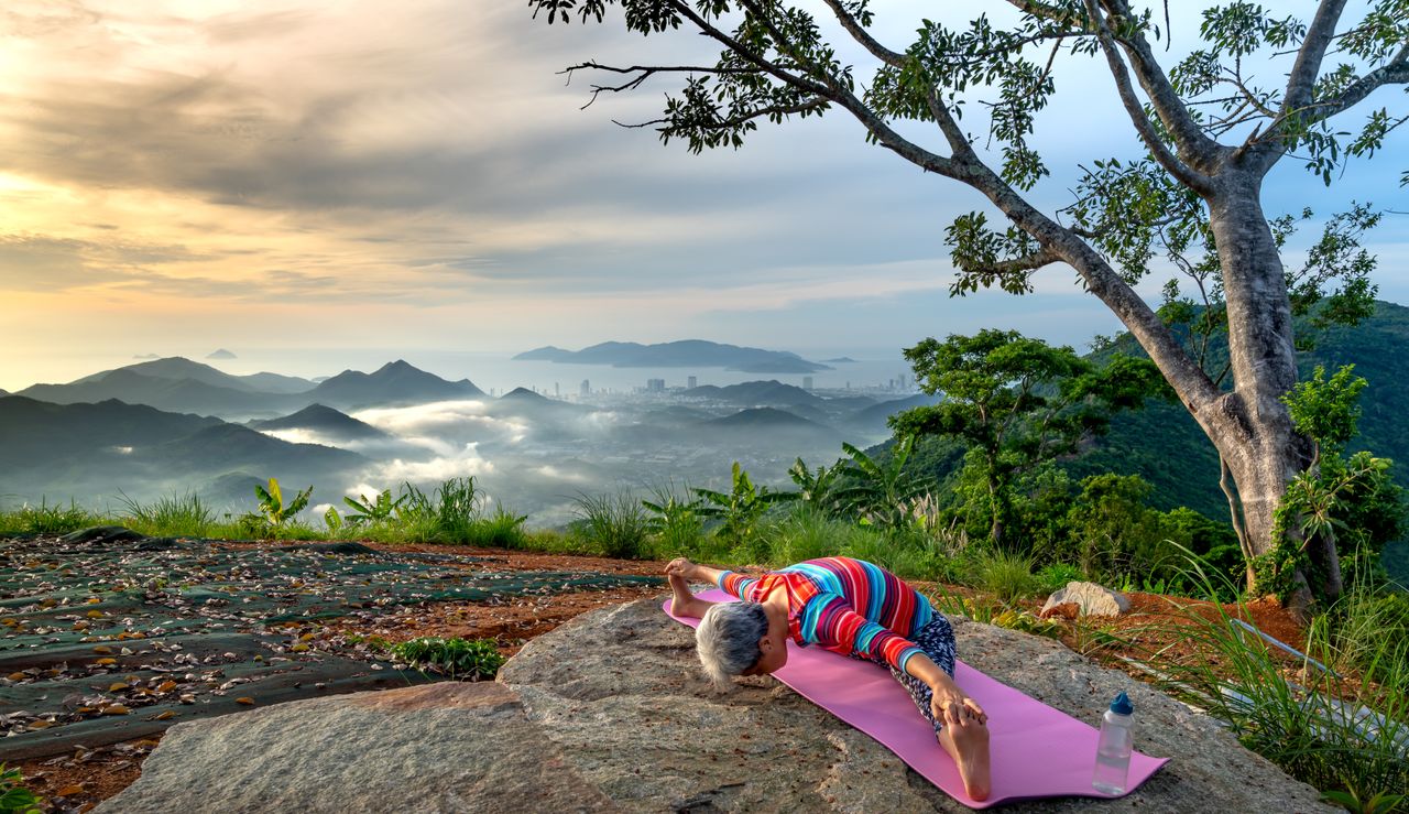 Home Senior woman doing yoga on a hilltop with a breathtaking mountain view near sunset.