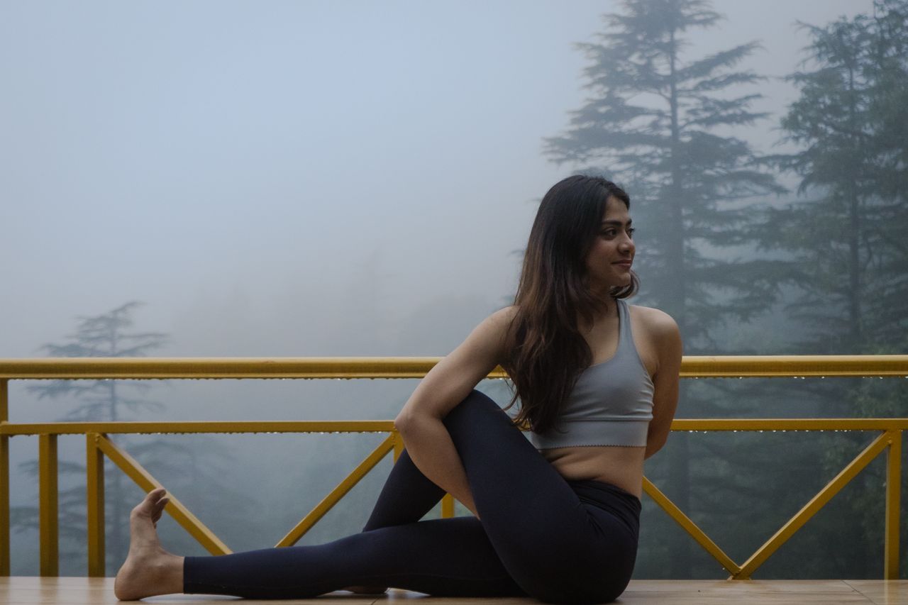Home Young woman doing yoga pose on a terrace in foggy Dharamshala, India, surrounded by pine trees.