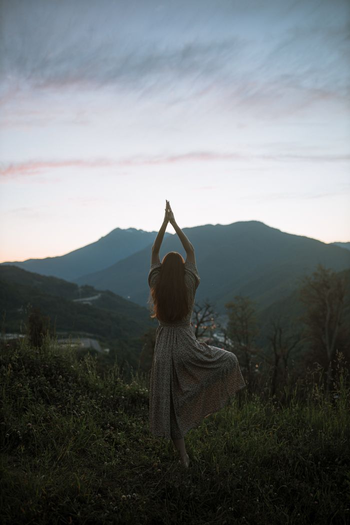 Home A woman in a dress performs tree pose against a mountainous sunrise backdrop, adding serenity and strength.