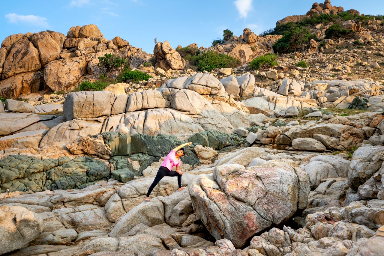 Home An Asian woman stretches on boulders in a scenic outdoor setting, perfect for travel and fitness themes.